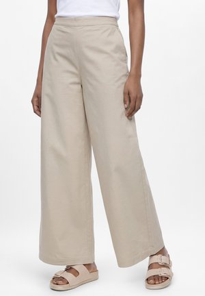 Beige weite Hose kombiniert mit beigen Sandalen mit Schnalle, getragen von einer Person vor einem einfarbigen weißen Hintergrund.