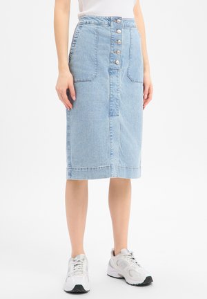 Frau, die einen hellblauen, knielangen Jeansrock mit großen Vorder Taschen und weißen Turnschuhen trägt und vor einem weißen Hintergrund steht.