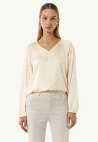 Hellrosa Bluse mit langen Ärmeln und V-Ausschnitt, glatter Textur und Raglanärmeln, kombiniert mit hellen, eng anliegenden Jeans.