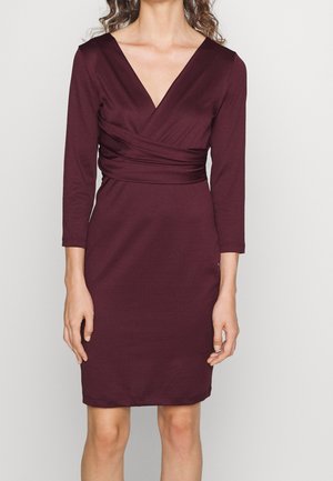 Vrouw draagt een nauwsluitende, knielange jurk in diep bordeauxrood met driekwartmouwen en een overslagtop met V-hals.