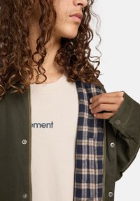 Grüne äußere Jacke mit Druckknöpfen und einem karierte Innenfutter. Helle beige T-Shirt mit blauer Schrift darunter. Lockige Haare sichtbar.