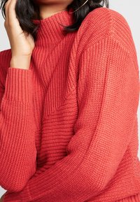 Pull en tricot rouge à col montant, texture côtelée et manches longues. Présente un motif tricoté diagonal sur la poitrine.