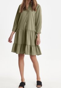 Sukienka w oliwkowym kolorze z guzikami, z długimi rękawami, warstwowym dołem z falbanami i fakturą w pionowe paski. Materiał swobodnie opada. Na nogach czarne klapki.