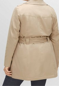 Beige verkkatakki, jossa on vyötärönauha, takana luhkakiila, nepparikiinnitys ja sileä kangaspinta. Viimeistelyyn kuuluu ompelusaumojen yksityiskohdat.