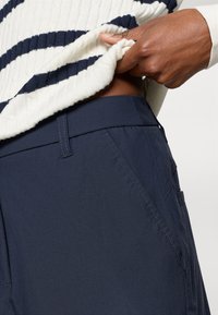 Navy blue bukser laget av lett materiale med flat front, sidelommer og subtile sømdetaljer. Livbåndet er litt synlig.