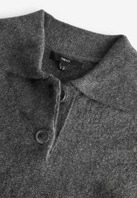 Pull en laine gris avec une finition texturée, présentant un col et deux boutons noirs sur le devant. Étiquette de la marque visible.