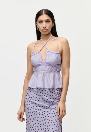 Mujer con cabello largo y oscuro que lleva un top halter lavanda con tirantes cruzados y una falda lavanda con lunares marrones, de pie frente a un fondo liso.