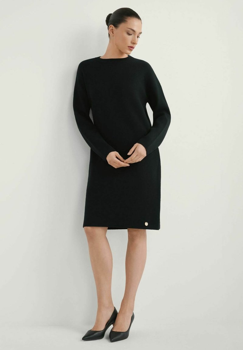 Kobieta w czarnej, długorękawowej sukience do kolan i czarnych szpilkach, stojąca z splecionymi rękami, patrząca w dół na bok.