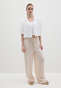 Weiße, kurz geschnittene Hemdbluse mit einer strukturierten Tasche, kombiniert mit fließenden beigen Hosen mit einem Kordelzugbund. Espadrille-Sandalen runden den Look ab.
