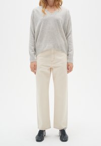Femme portant un pull gris clair à col en V, un pantalon large crème et des chaussures noires à lacets, debout devant un fond blanc.