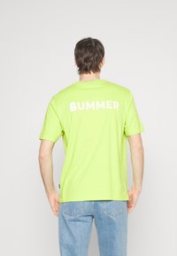 Personne de dos portant un T-shirt vert citron avec "BUMMER" en blanc et un jean clair sur fond blanc.