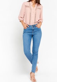 Kvinna som bär en ljusrosa blus med spetsdetaljer och blå skinny jeans, stående med händerna i fickorna och med korslagda fötter.