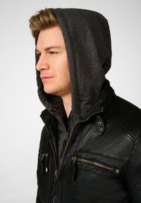 Schwarze Lederjacke mit einer dunkelgrauen Kapuzeninnenschicht. Auffällige Details sind die Reißverschlusstaschen und die gerippten Schulterakzente.