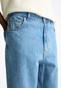 Los vaqueros de denim azul claro presentan una cintura alta, un diseño de cinco bolsillos y detalles de costura sutiles, con una textura ligeramente desgastada.