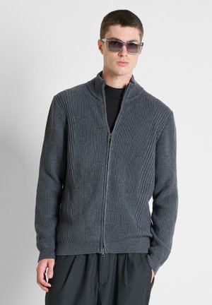 Junger Mann, der einen grauen, gerippten Reißverschluss-Pullover, schwarze Hose, schwarzes Hemd und graue Sonnenbrille trägt, steht vor einem einfachen hellen Hintergrund.
