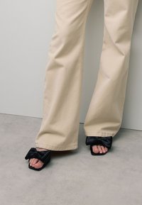 Zwarte sandalen met een vierkante neus, voorzien van een satijnen strik op de band. Gepaarde met loszittende beige broeken, op een gestructureerde grijze vloer.