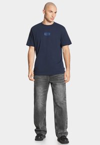 Granatowa bawełniana koszulka z małym niebieskim logo na piersi, w połączeniu z szarymi dżinsami i czarnymi sneakersami. Luźny krój i styl.