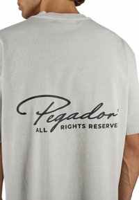 Grå bomulds t-shirt med et sort trykt logo "Pegador" og teksten "ALLE RETTIGHEDER RESERVERET!" på ryggen. Rund hals og korte ærmer.