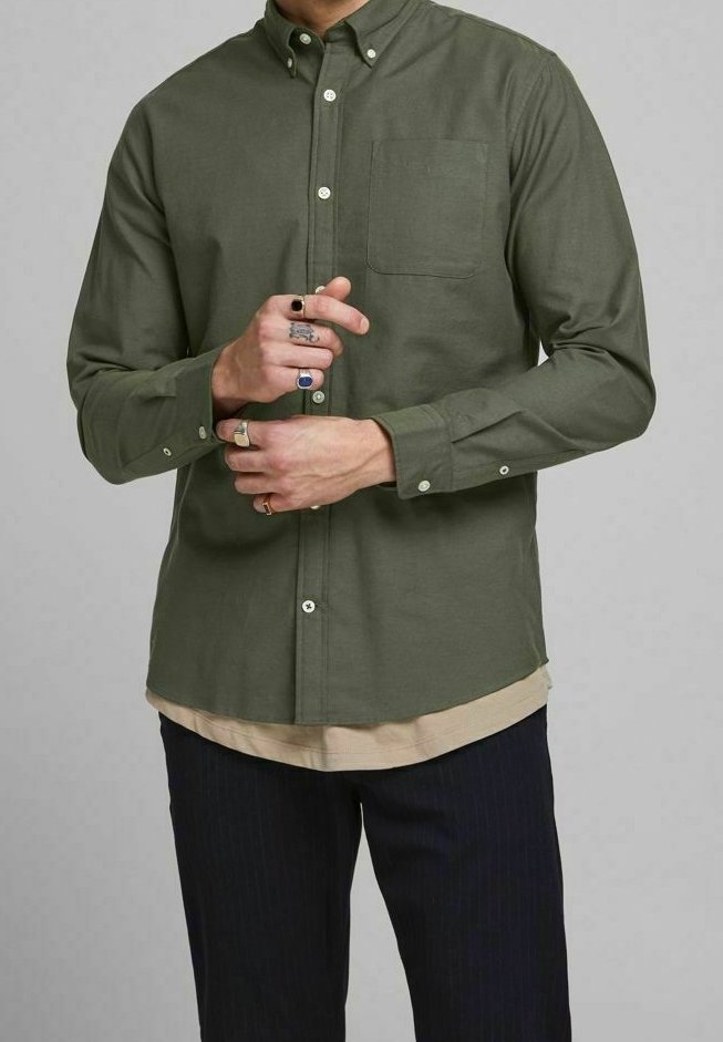 Green button-up shirt with a collar, chest pocket, and long sleeves. Worn over a beige layered top and paired with dark pinstriped pants.