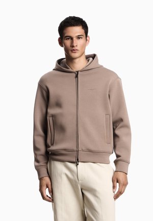 Felpa beige con zip realizzata in materiale morbido, dotata di cappuccio con coulisse, due tasche frontali e polsini a coste. Design più pulito con accenti minimi.