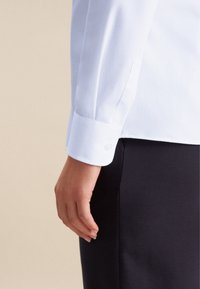 Witte lange mouwen overhemd met een knoopsluiting bij de manchetten, gladde stof, getailleerde pasvorm en subtiele glans, gepresenteerd naast een donkere broek tegen een neutrale achtergrond.