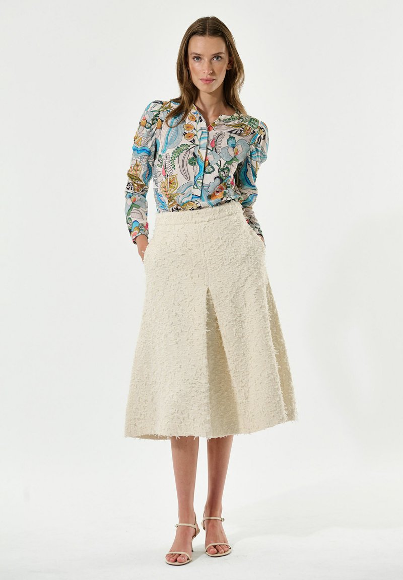 Bunte gemusterte Bluse mit langen Ärmeln, kombiniert mit einem strukturierten cremfarbenen Rock mit einem Schlitz vorne und Taschen. Einfache beige Absätze.