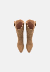 Bottines en suede couleur tan allant jusqu'au genou, avec des pointes effilées, des coutures décoratives et des languettes latérales. Texture lisse et détails matériels minimaux.