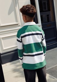 Zeleno-bíle pruhovaná košile s dlouhým rukávem a námořnickými detaily, s límečkem, v kombinaci s tmavými džínami. Viditelná textura látky.