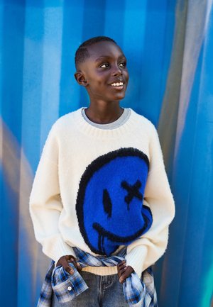 Cream sweater with a large blue smiley face design, ribbed neckline, and soft texture. Paired with blue plaid shirt tied at the waist.
