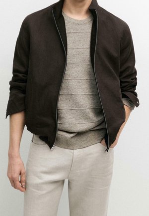 Homme portant une veste zippée marron foncé sur un pull en maille côtelée beige et un pantalon beige clair, une main dans la poche, sur un fond uni.