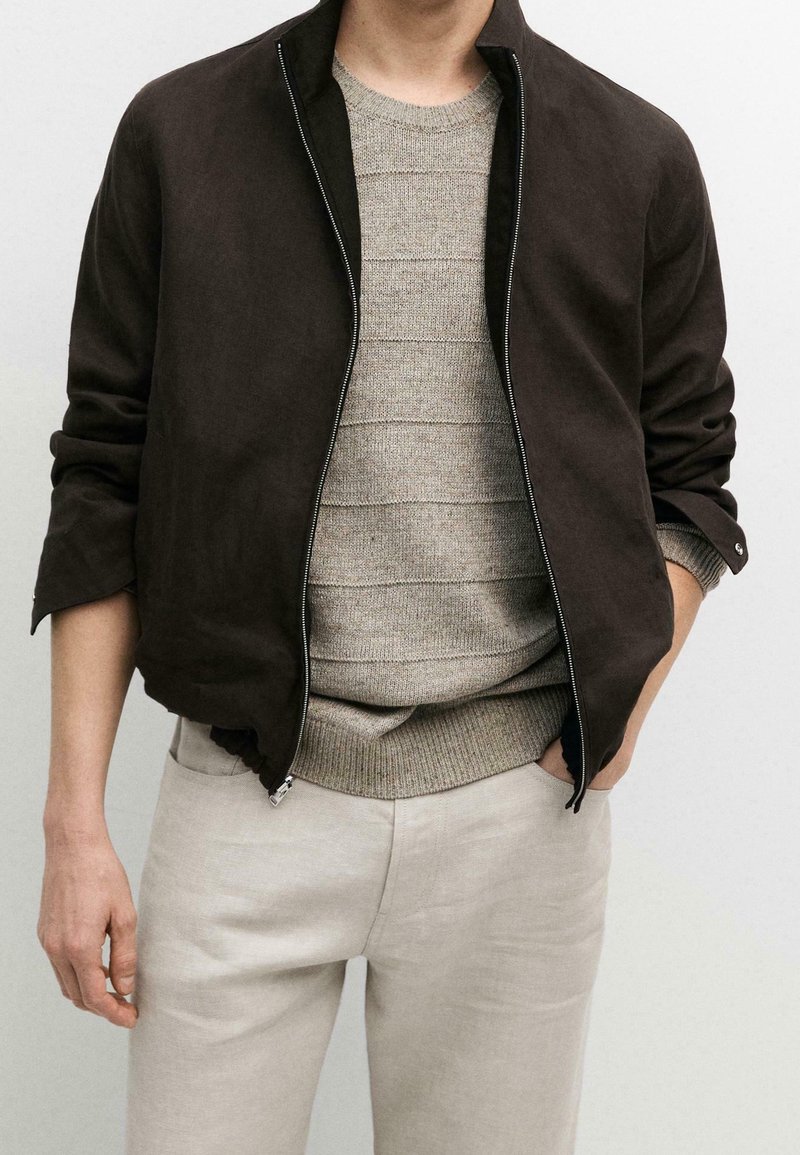 Homme portant une veste zippée marron foncé sur un pull en maille côtelée beige et un pantalon beige clair, une main dans la poche, sur un fond uni.