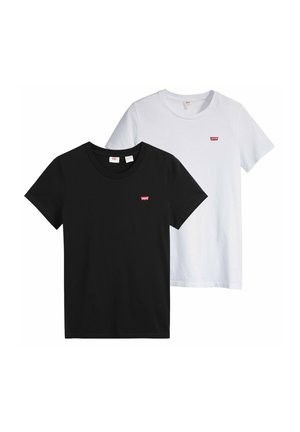 Giovane donna con lunghi capelli castani che indossa una t-shirt nera Levi's e jeans scuri su sfondo grigio.