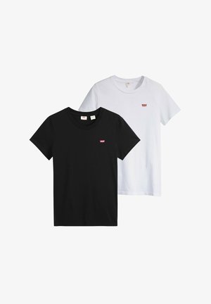 Jeune femme aux longs cheveux bruns portant un t-shirt noir Levi's et un jean foncé sur un fond gris.