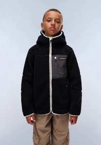 Fant s črnim flis jakno z zadrgo na prsnem žepu in bež cargo hlačami, ki stoji pred enobarvno svetlo ozadje.