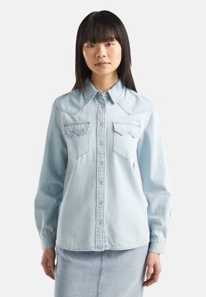 Jonge vrouw met stijl zwart haar, gekleed in een lichtblauw denimblouse met knopen en een bijpassende denimrok tegen een effen achtergrond.