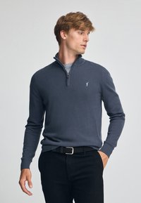 Grå zip-up tröja i mjukt tyg, med ståkrage och en broderad logotyp på vänster bröst. Bärs med svarta byxor.