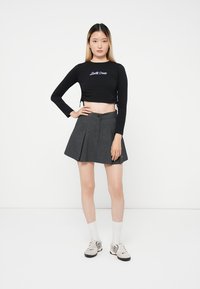 Young woman stands with hand on hip wearing black long-sleeve crop top, gray pleated skirt, white socks, and sneakers on white background.