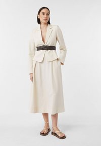 Cremfarbener Leinenblazer mit tailliertem Design, kombiniert mit einem passenden Midikleid und einem strukturierten dunklen Gürtel. Stylisch ergänzt mit Sandalen mit goldenen Riemen.