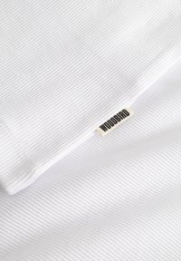 Tissu blanc à côtes plié avec une petite étiquette beige affichant le texte « WOODBIRD » en lettres noires.