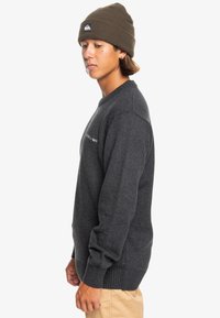 Grå stickad tröja med ribbstickade muddar och fåll. Har en logga på övre vänstra sidan. Matchas med en brun mössa. En enkel, minimalistisk design.