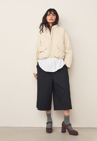 Beige bomberjacka med två frampickar, överlagrad på en vit skjorta. Bärs med svarta vida byxor och bruna klackskor, kombinerat med grå strumpor.