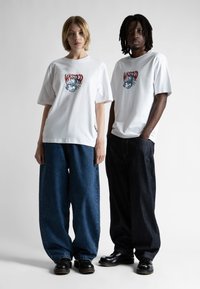 Bílé trička s grafickým potiskem, kombinovaná s volnými modrými a tmavě modrými džíny. Černé boty dotvářejí outfity.