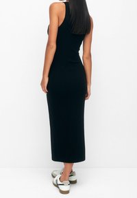 Femme aux longs cheveux bruns portant une robe mi-longue noire sans manches et des baskets blanches, vue de dos sur un fond blanc uni.