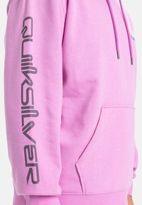 Ružová mikina s predným vreckom a šnúrkou. Obsahuje veľké, tmavošedé logo "Quiksilver" pozdĺž ľavého rukáva. Mäkká textúra tkaniny.