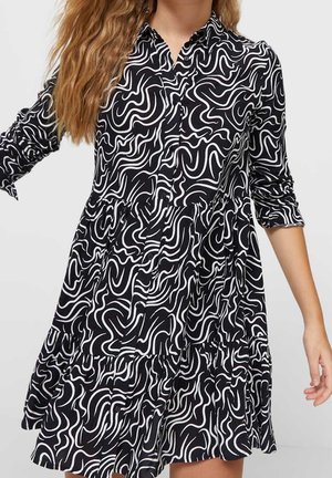 Femme portant une robe noire avec des lignes blanches abstraites ondulées, manches longues retroussées, devant boutonné, et jupe à volants.
