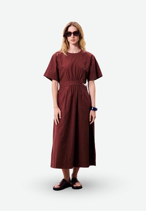 Femme aux longs cheveux blonds portant une robe midi couleur bordeaux, des lunettes de soleil foncées, des sandales noires et un bracelet sombre, debout devant un fond blanc.