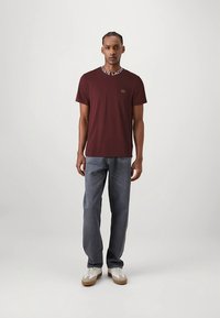 Tamsiai raudonas apvalios iškirptės marškinėlis su baltu logotipu ant apykaklės, derinamas su mėlynomis džinsais ir baltais sportbačiais su smėlio spalvos padais, dėvimas modelio.