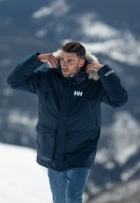 Námořnická modrá zateplená parka s kapucí lemovanou kožešinou, několika zipovými kapsami a logem HH na hrudi; hladká textura vhodná pro chladné počasí.