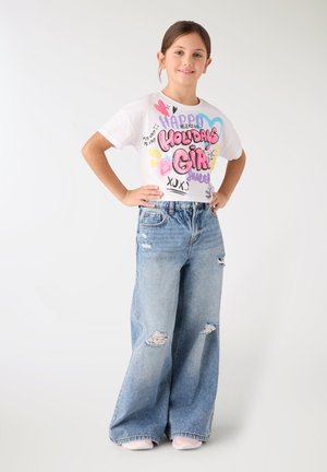 Ragazza giovane che sorride con le mani sui fianchi, indossando un top corto bianco con stampa "Holidays Girl" e jeans blu a gamba larga strappati.