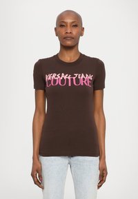 Brauns īsām piedurknēm T-krekls, izgatavots no kokvilnas, ar rozā tekstu "VERSACE JEANS COUTURE" uz krūtīm. Pielāgots dizains ar apaļu kakla izgriezumu.